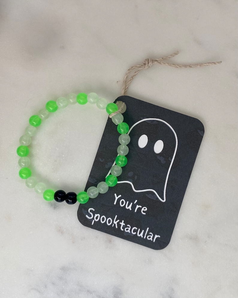 K&ouml;nnte beinhalten: Ein Armband mit abwechselnd gr&uuml;nen und durchsichtigen Perlen und zwei schwarzen Perlen. Ein schwarzes Etikett mit einer Geisterillustration und dem Schriftzug "You're Spooktacular" ist mit Bindfaden am Armband befestigt. Das Armband liegt auf einer wei&szlig;en Oberfl&auml;che.