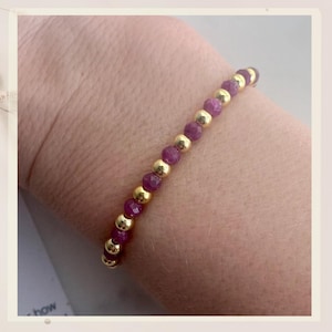 May include: A gold and pink beaded bracelet with small, faceted beads. The bracelet is on a person's wrist.