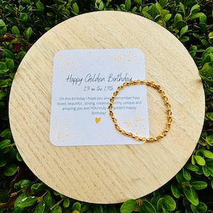 May include: A gold bracelet with amber beads and a card that reads "Happy Golden Birthday 13 on the 13th" with a gold heart. The card has a white background with gold glitter.