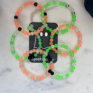 K&ouml;nnte beinhalten: Mehrere Armb&auml;nder mit Perlen in Orange, Gr&uuml;n und transparent. Einige Armb&auml;nder haben schwarze Perlen. Ein Anh&auml;nger mit einer Geistergrafik und dem Text "spooktacular" ist befestigt. Die Armb&auml;nder liegen auf einer wei&szlig;en Marmoroberfl&auml;che.