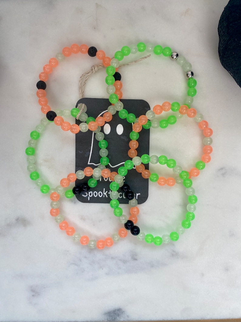K&ouml;nnte beinhalten: Mehrere Armb&auml;nder mit Perlen in Orange, Gr&uuml;n und transparent. Einige Armb&auml;nder haben schwarze Perlen. Ein Anh&auml;nger mit einer Geistergrafik und dem Text "spooktacular" ist befestigt. Die Armb&auml;nder liegen auf einer wei&szlig;en Marmoroberfl&auml;che.
