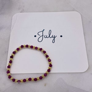 May include: A purple and gold beaded bracelet with the word "July" written on a white card.