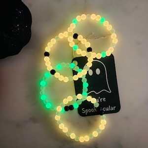 K&ouml;nnte beinhalten: Leuchtende Perlenarmb&auml;nder mit Halloween-Motiv. Die Armb&auml;nder haben leuchtende Perlen in Wei&szlig;, Gr&uuml;n und Schwarz. Ein Anh&auml;nger mit einer Geisterillustration und dem Schriftzug "you're Spooktacular" ist angebracht.