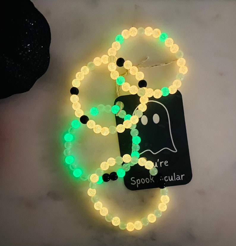 K&ouml;nnte beinhalten: Leuchtende Perlenarmb&auml;nder mit Halloween-Motiv. Die Armb&auml;nder haben leuchtende Perlen in Wei&szlig;, Gr&uuml;n und Schwarz. Ein Anh&auml;nger mit einer Geisterillustration und dem Schriftzug "you're Spooktacular" ist angebracht.