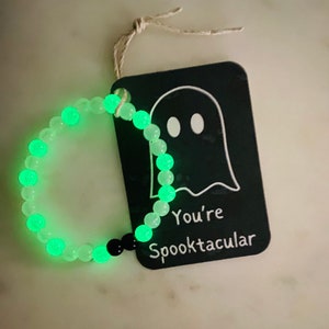 K&ouml;nnte beinhalten: Ein Armband mit leuchtend gr&uuml;nen Perlen und einer schwarzen Perle. Ein schwarzes Etikett mit einer wei&szlig;en Geisterillustration und dem Text "You're Spooktacular" ist mit Bindfaden befestigt. Halloween-Themen-Accessoire.
