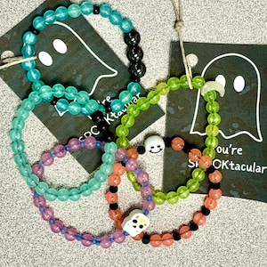 K&ouml;nnte beinhalten: Eine Sammlung von Armb&auml;ndern mit Perlen in verschiedenen Farben, darunter T&uuml;rkis, Gr&uuml;n, Lila und Orange. Einige Armb&auml;nder haben schwarze Perlen und wei&szlig;e Totenkopf-Anh&auml;nger. Jedes Armband ist auf einer Schnur aufgef&auml;delt.