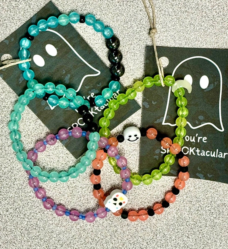 K&ouml;nnte beinhalten: Eine Sammlung von Armb&auml;ndern mit Perlen in verschiedenen Farben, darunter T&uuml;rkis, Gr&uuml;n, Lila und Orange. Einige Armb&auml;nder haben schwarze Perlen und wei&szlig;e Totenkopf-Anh&auml;nger. Jedes Armband ist auf einer Schnur aufgef&auml;delt.