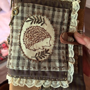 May include: A fabric-covered journal featuring a brown and tan plaid pattern with a central oval patch embroidered with a hedgehog and leaf motifs. The cover is accented with cream-coloured lace trim, visible hand-stitching, and metal charms.