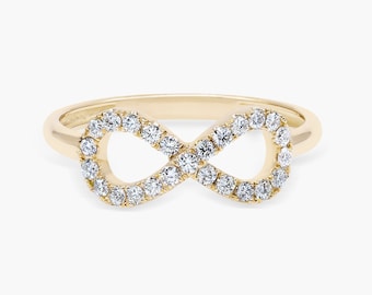 Könnte beinhalten: Ein goldfarbener Ring mit einem Unendlichkeitszeichen-Design. Das Unendlichkeitszeichen ist mit zahlreichen kleinen, runden, klaren Edelsteinen besetzt. Das Ringband ist aus schlichtem Gold. Der Ring ist vor einem weißen Hintergrund platziert. Der Ring ist etwa 2,5 cm breit.