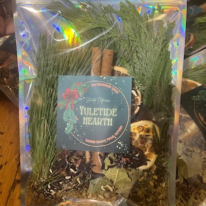 Puede incluir: Una bolsa holográfica transparente llena de hierbas secas, especias y productos botánicos. La bolsa contiene agujas de pino verdes, palitos de canela, rodajas de naranja secas y una etiqueta que dice "Yuletide Hearth". La etiqueta también incluye el texto "Sacred Roots from Saturn!"