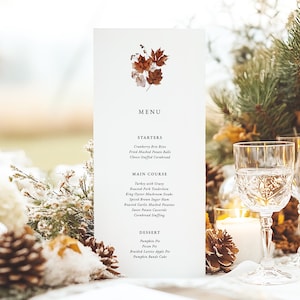 May include: A white menu card with a brown leaf design, listing a Thanksgiving dinner menu with starters, main course, and dessert options. The menu is set on a white tablecloth with pine branches, pine cones, and white candles.
