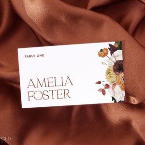 Op de afbeelding: Een witte tafelkaart met de tekst "Table One" en "Amelia Foster" in een modern lettertype. De kaart is versierd met een bloemenillustratie met bordeauxrode, oranje en gele bloemen.