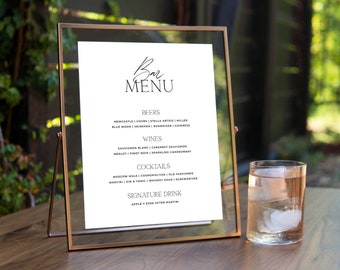 Puede incluir: Un cartel de menú de bar blanco con marco dorado, que enumera cervezas, vinos, cócteles y una bebida de autor. El cartel está sobre una mesa de madera con un vaso de agua con hielo.