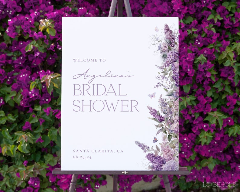 May include: A white welcome sign with purple text that reads 'Welcome to Angelina's Bridal Shower' and 'Santa Clarita, CA 06.24.24'. The sign is decorated with a watercolor illustration of purple flowers and butterflies on the right side.