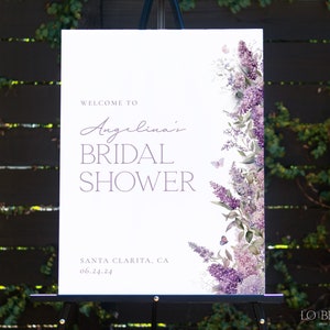 May include: A white bridal shower sign with purple flowers and butterflies. The sign reads 'Welcome to Angelina's Bridal Shower' and 'Santa Clarita, CA 06.24.24'.