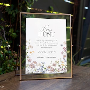 May include: A white sign with black text and floral illustrations announcing a ring hunt game at a bridal shower. The text reads "Ring Hunt" and "There are rings hidden throughout the shower, the one who finds the most rings by the time the first gift is unwrapped wins a special prize! Good Luck" and "Kimora's Bridal Shower".