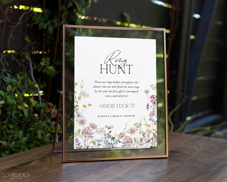May include: A white sign with black text and floral illustrations announcing a ring hunt game at a bridal shower. The text reads "Ring Hunt" and "There are rings hidden throughout the shower, the one who finds the most rings by the time the first gift is unwrapped wins a special prize! Good Luck" and "Kimora's Bridal Shower".