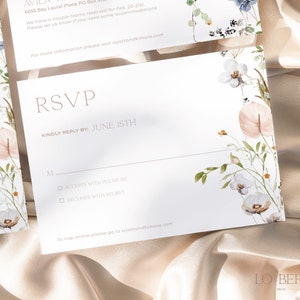 May include: RSVP card with a floral design. The card has a white background with a pink and white floral pattern. The text "RSVP" is in a light brown font. The text "KINDLY REPLY BY: JUNE 15TH" is in a light brown font. The text "M" is in a light brown font. The text "ACCEPTS WITH PLEASURE" is in a light brown font. The text "DECLINES WITH REGRET" is in a light brown font. The text "To RSVP online please go to wyattandkimora.com" is in a light brown font.
