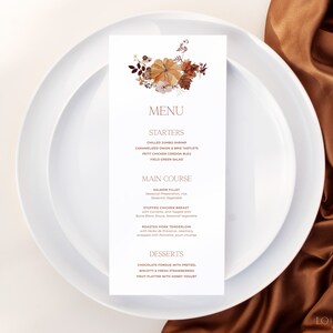 Op de afbeelding: Een witte menukaart met een herfstthema bloemenontwerp in tinten oranje, bruin en wit. Het menu bevat een selectie van voorgerechten, hoofdgerechten en desserts. De tekst op de menukaart luidt: "MENU, STARTERS, CHILLED JUMBO SHRIMP, CARAMELIZED ONION & BRIETARTLETS, PETIT CHICKEN COOCH BLEU, FIELD GREEN SALAD, MAIN COURSE, SALMON FILLET, STUFFED CHICKEN BREAST, BOASTED FORK TENDERLOIN, DESSERTS, CHOCOLATE FONDUE WITH PRETZEL, BISCOTTI & FESTRAWBER, FRUIT PLATTER WITH HONEY YOGURT, LO BEHOLD."