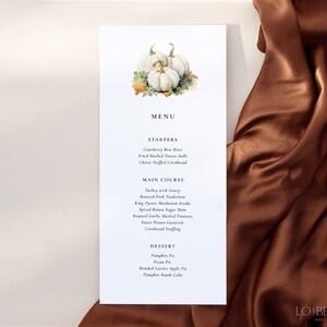 May include: A white card with a watercolor illustration of white pumpkins and fall leaves. The card has a menu for a Thanksgiving dinner with starters, main course, and dessert options. The text on the card reads "MENU", "STARTERS", "MAIN COURSE", "DESSERT", "Cranberry Brie Bites", "Fried Mashed Potato Balls", "Cheese Stuffed Cornbread", "Turkey with Gravy", "Roasted Pork Tenderloin", "King Oyster Mushroom Steaks", "Spiced Brown Sugar Ham", "Roasted Garlic Mashed Potatoes", "Sweet Potato Casserole", "Cornbread Stuffing", "Pumpkin Pie", "Pecan Pie", "Braided Lattice Apple Pie", "Pumpkin Bundt Cake", and "LO BEHOLD".