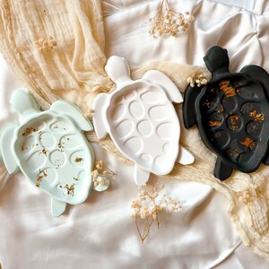 Könnte beinhalten: Drei Keramik-Schalen in Form von Schildkröten. Eine ist weiß, eine hellblau und eine schwarz. Jede Schale hat goldene Flocken auf der Oberfläche.