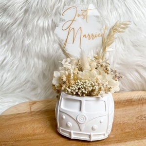 Könnte beinhalten: Weißer Keramik-VW-Bus-Pflanztopf gefüllt mit getrockneten weißen Blumen und Grün. Ein klares Acrylschild mit goldenen Buchstaben lautet "Just Married".