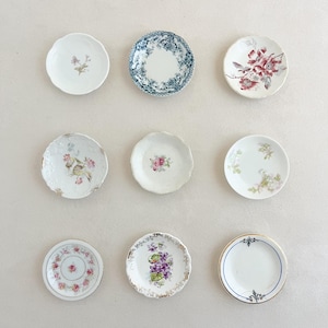 May include: A collection of nine small, round ceramic saucers arranged in a grid. Each saucer features a unique decorative pattern, including floral motifs in pink, purple, and green, a blue and white transferware design, and gold-trimmed edges.