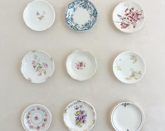 May include: A collection of nine small, round ceramic saucers arranged in a grid. Each saucer features a unique decorative pattern, including floral motifs in pink, purple, and green, a blue and white transferware design, and gold-trimmed edges.