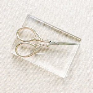 May include: A pair of gold-toned, ornate scissors with a silver blade on a clear acrylic surface.