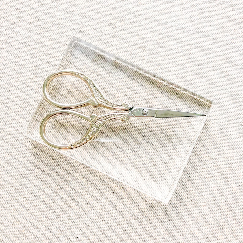 May include: A pair of gold-toned, ornate scissors with a silver blade on a clear acrylic surface.