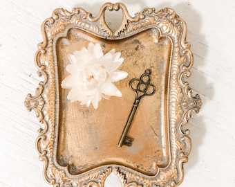 May include: A rectangular gold-toned metal tray with an ornate, raised border featuring intricate scrollwork. Resting on the tray is a single antique-style metal key with a decorative bow and a white, multi-petaled dried flower blossom.