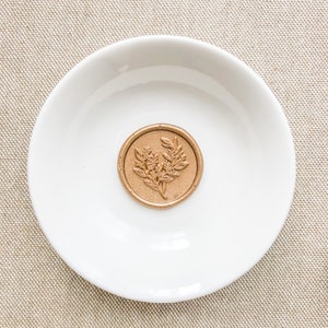 May include: A gold wax seal with a floral design on a white plate.