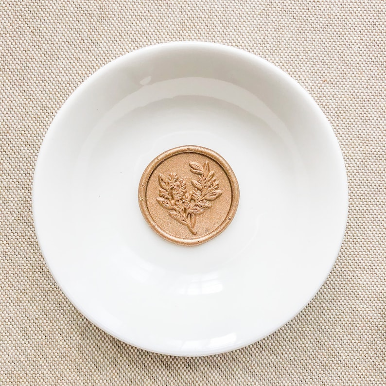 May include: A gold wax seal with a floral design on a white plate.