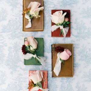 May include: Five small vintage books with pink roses and greenery tied with white ribbon on top of each book. The books are arranged vertically on a blue and white patterned surface.
