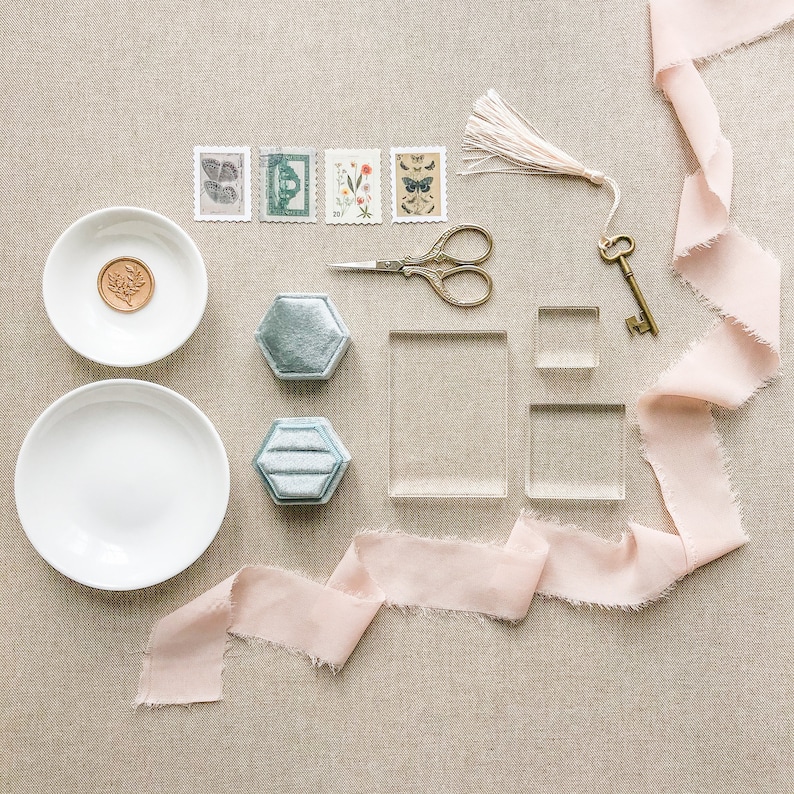 May include: A flat lay of wedding stationery supplies on a beige linen background. The items include two white bowls, two blue velvet ring boxes, two clear acrylic boxes, a pair of antique scissors, a vintage key, a pink ribbon, and four vintage postage stamps.