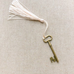 May include: An antique-style key with a decorative tassel attached to the end. The key is made of brass and has a worn, aged look. The tassel is made of a soft, white material.