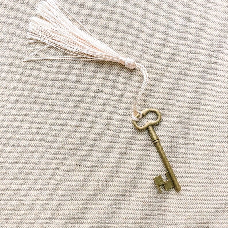 May include: An antique-style key with a decorative tassel attached to the end. The key is made of brass and has a worn, aged look. The tassel is made of a soft, white material.