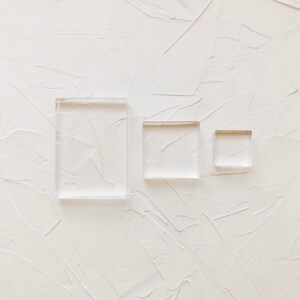 May include: Three clear acrylic stamps of different sizes, arranged on a white textured background.