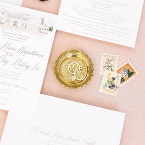 May include: A wedding invitation suite with a gold ring dish holding two rings, invitation cards, and floral stamps. The invitation cards have elegant script and a watercolor illustration of a building. The background is a soft pink fabric.