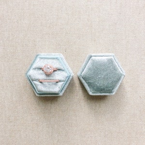 May include: A light blue velvet ring box with a hexagon shape. The box is open and contains a diamond engagement ring and a wedding band. The rings are set in rose gold.