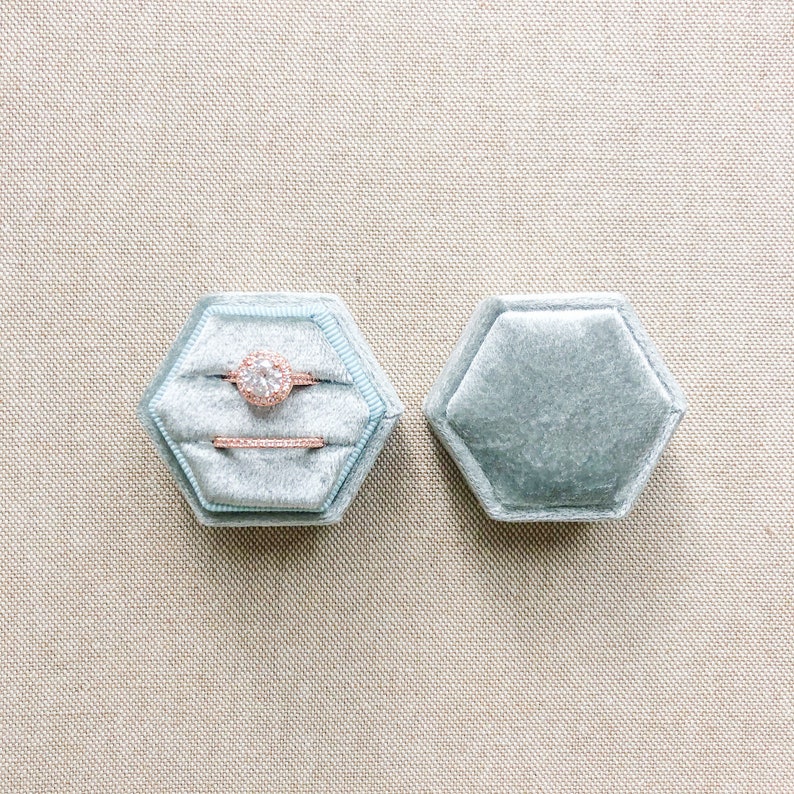 May include: A light blue velvet ring box with a hexagon shape. The box is open and contains a diamond engagement ring and a wedding band. The rings are set in rose gold.