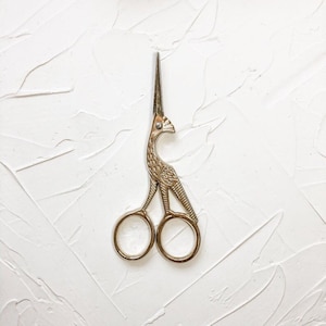May include: Gold-toned bird-shaped scissors with ornate details. The scissors have two round handles.