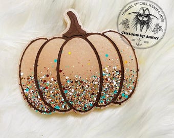 May include: A pumpkin-shaped ornament featuring a tan base with dark brown outlines and a dark brown stem. The lower portion is decorated with a dense layer of multi-colored glitter in shades of teal, copper, and white. A circular logo reads Customs by Amber.
