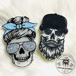 May include: Two skull-shaped ornaments. The left skull features a messy bun with a light blue glitter headband and silver glitter aviator sunglasses. The right skull wears a black baseball cap and has a detailed black beard and mustache.