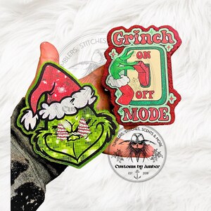 May include: Two glittery, die-cut decorative ornaments featuring a green character with a red Santa hat and a light switch graphic with the text "GRINCH ON OFF MODE". The ornaments have a textured, sparkling finish and vibrant red, green, and white colors.