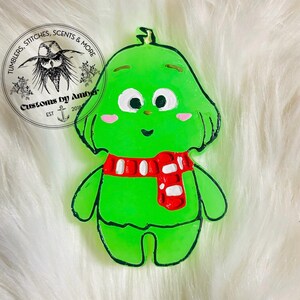 May include: A bright green character-shaped ornament with a red and white scarf, large white eyes, and pink cheeks. The figure has a smooth, glossy finish with black outlines. A circular logo in the corner reads Tumblers, Stitches & More Customs by Amber.