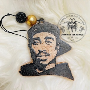 May include: A hanging ornament featuring a portrait of Tupac Shakur in black and tan, attached to a black string with one black textured bead and one gold metallic bead. A circular logo with the text Tumblers, Stitches, Scents & More is visible.