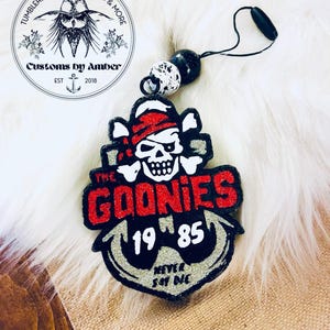 May include: A black, red, and silver glittery hanging ornament featuring a skull wearing a red bandana, the text THE GOONIES, the numbers 19 85, and the phrase NEVER SAY DIE, with a black anchor graphic and a black cord with two beads.