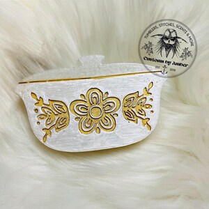 May include: A white, oval-shaped lidded container featuring a gold-colored floral pattern on the front. The design includes a central five-petaled flower flanked by two leafy sprigs. A circular logo with the text Tumblers, Scents & More is visible.