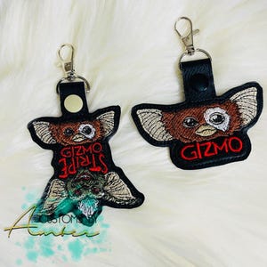 May include: Two black leather keychains with silver metal lobster clasps. One keychain features embroidered faces of Gizmo and Stripe with red text. The other keychain features an embroidered face of Gizmo with the red text GIZMO.